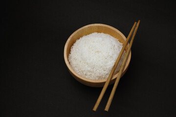 Wooden bowl filled with freshly steamed white rice with chopsticks on top, isolated on black background for minimalist food or culinary theme.