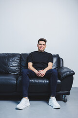 A young handsome man in a black t-shirt is sitting on a black sofa. A man rests sitting on a comfortable sofa.