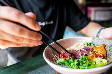 close-up hands using chopsticks to eat vibrant bowl of Asian noodles with vegetables and meat. image highlights fresh ingredients and colorful presentation, perfect for culinary and lifestyle themes.