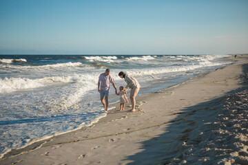 A young perents holds her little daughter by the hand and together they walk along the ocean towards the sunset.