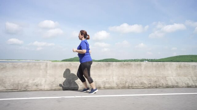A side view of a middle-aged, overweight Asian woman jogging for exercise on a scenic road by a reservoir, aiming for better health and weight loss