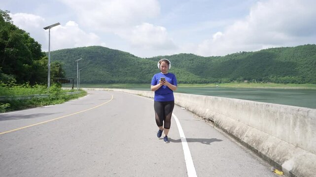 A middle-aged, overweight Asian woman wearing headphones is walking for exercise on a road while using a smartphone, aiming for better health and weight loss, and relaxing during her workout