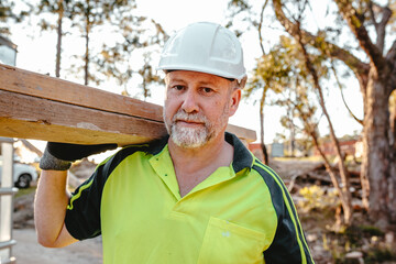 A man wearing hardhat carrying a stack of lumber on his shoulder at a construction site.