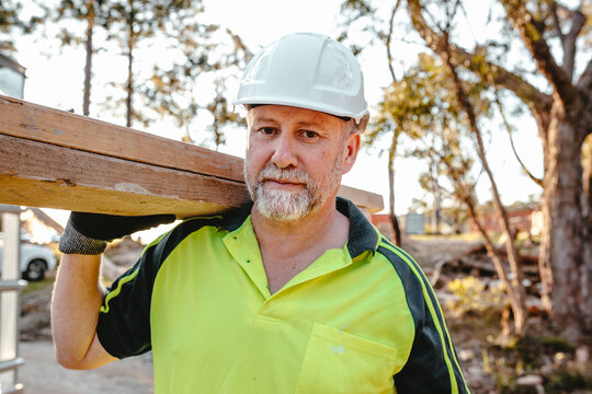 A man wearing hardhat carrying a stack of lumber on his shoulder at a construction site.