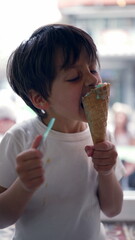 Young boy eating ice cream with a spoon and cone in hand, enjoying a sweet treat indoors,...