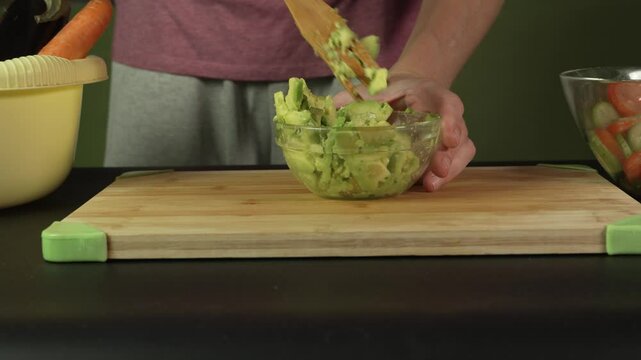 Hands of middle man european mashes avocado in glass bowl with wooden spatula for guacamole dip, front side view, medium shot. Healthy lifestyle, vegetarian or vegan, raw food diet, cooking at home