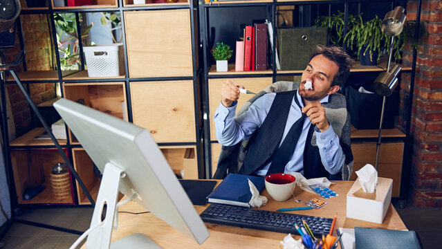 Sick businessman sits wrapped in blanket at desk, drinking from large cup. Instead of taking day off, he working while clearly unwell, affecting productivity. Concept of daily work challenges.