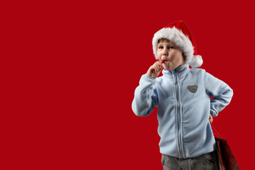 Child in Santa Claus hat with a lollipop in his mouth and with carrier bag in his hand on red background. Copy space.