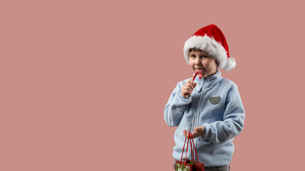 A little girl in a Santa Claus hat is licking a cane. She has a paper gift bag in her other hand. The background is pink. Copy space.