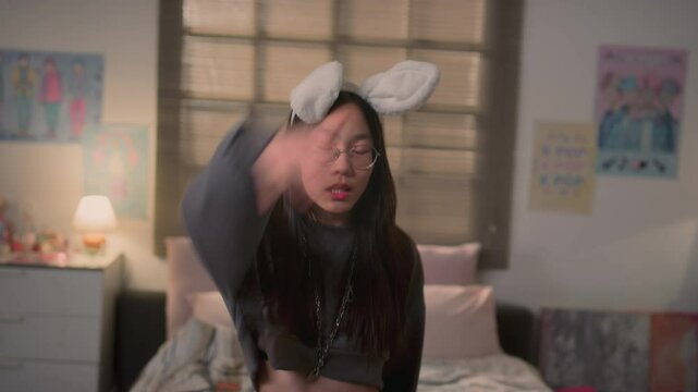 Long-haired Asian girl energetically dancing to k-pop music, lip-syncing and looking at camera in bedroom