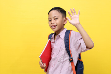 Happy asian schoolboy standing while raise hand, carrying a book and school bag . Isolated on yellow