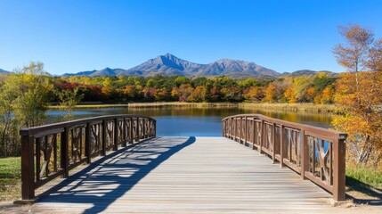 Obraz premium A wooden bridge welcomes visitors to a serene lake, framed by autumn foliage in red and orange, under clear blue skies and distant mountains, evoking tranquility