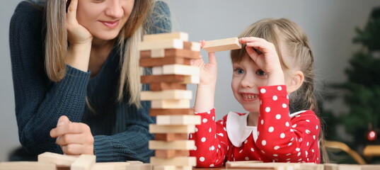 Portrait of pretty little kid pulling wooden block out of tower and smile. Mother join daughter in...