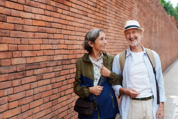 Senior Caucasian couple strolling along ancient brick wall in Chiang Mai, Thailand. Man wearing hat and striped shirt, woman in casual jacket. Both laughing and enjoying a leisurely day. urban travel.