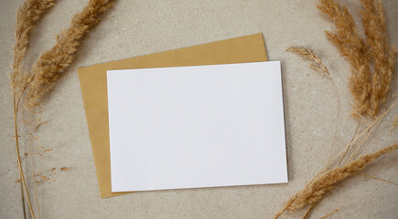 Blank white horizontal greeting card, golden envelope and dry grass on beige textured background. Top view flat lay with copy space. Greeting card mock up. © KatuSka