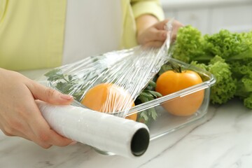 Woman putting plastic food wrap over glass container with fresh vegetables at white marble table,...