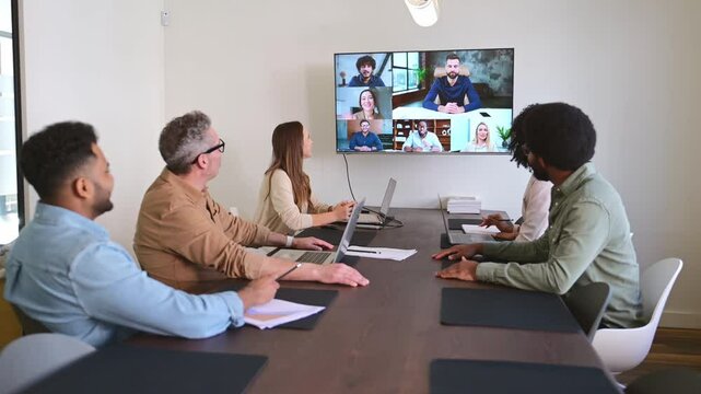 Team of professionals sits around conference table, engaging in an online video conference with remote colleagues shown on a large screen. The integration of digital tools in modern work environments