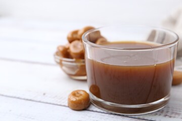 Tasty caramel sauce in bowl and candies on white wooden table, closeup