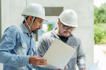 Asian male engineers reviewing construction plans outdoors. wearing safety helmets, one holding blueprints and reviewing details on tablet. environment features concrete structures under construction