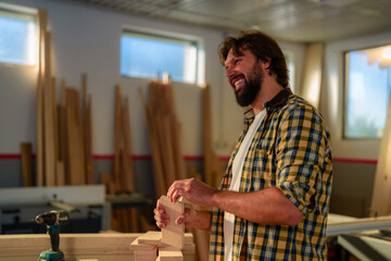 Portrait of young bearded smiling woodworker who sanding a piece of wood in workshop.