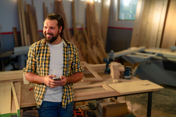 A carpenter in a workshop looking at his smartphone while leaning against a workbench.