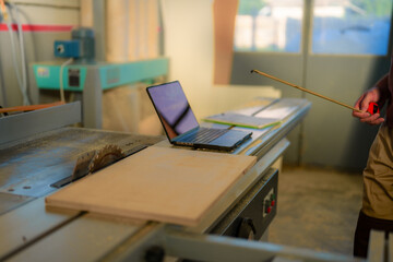 A person measuring a wooden board with a tape measure next to a laptop in a workshop.