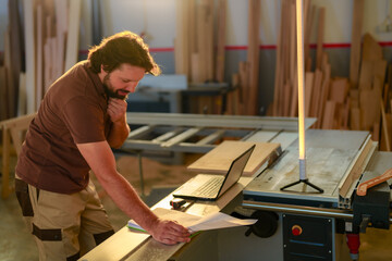 Craftsman using laptop for reviewing the project he work on in woodworking workshop.
