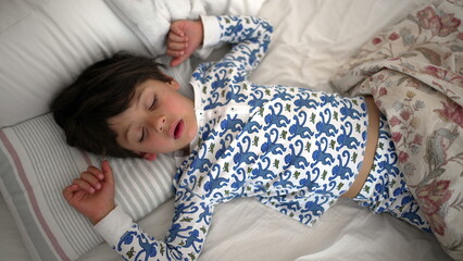 A young child is sleeping peacefully with arms stretched, in printed pajamas, covered partially with a floral blanket