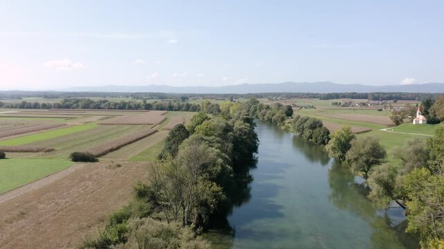 Flying Above crystal clear emerald river Kolpa flowing through green farmland and wooded areas with distant mountains. Perfect for projects focused on agriculture, rural landscapes, natural beauty.