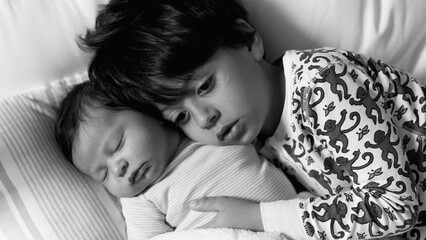 Black and white of older brother resting his head on his newborn sibling, both lying together on a...