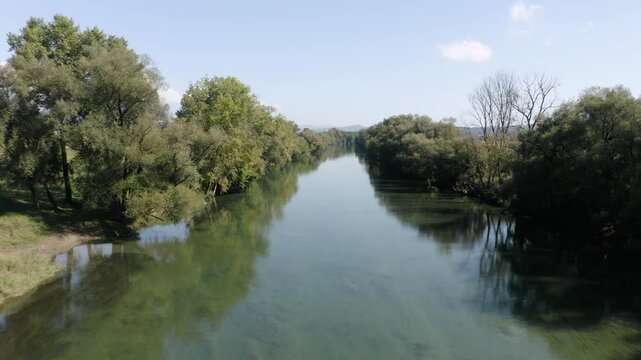 Flying over straight, calm river Kolpa reflecting a bright blue sky. The river is bordered by dense trees on both sides, casting shadows on the clear water. A peaceful nature scene.