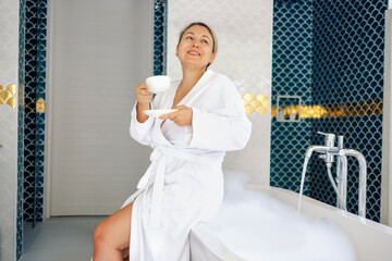 Young woman in white bathrobe drinks coffee and takes bubble bath. Blonde is smiling and enjoying hot fragrant drink. Lady is getting ready to take bath and relax at home.