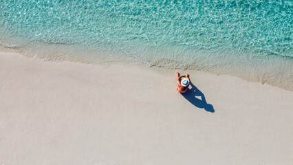 An aerial view of a young man in a white hat sitting on the ocean shore. A man enjoys the seascape. A man is relaxing on a clean sandy beach.
