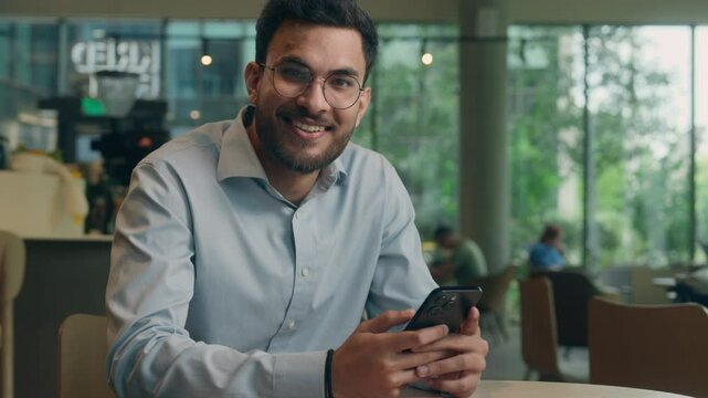 Indian Arabian business man male happy traveler tourist client guest visitor in cafe restaurant positive smiling ethnic guy businessman using mobile phone app online cellphone cheerful smile at camera