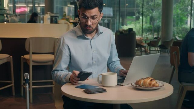 Indian man Arabian businessman guy male freelancer business worker customer visitor at cafe table online working laptop computer chatting using devices mobile phone read message email typing on pc