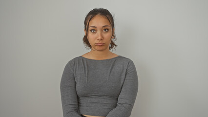 A young hispanic woman stands against a white isolated background, looking pensive with a neutral...