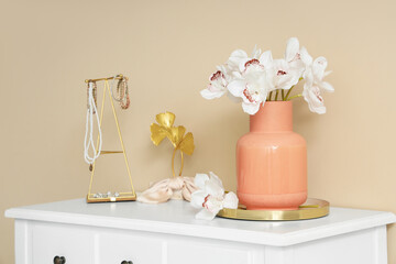 Vase with beautiful orchid flowers and jewelry on dressing table near beige wall