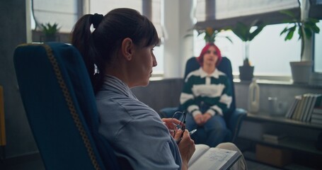 Back view of female therapist with notebook sitting in chair, providing therapy for teenager....
