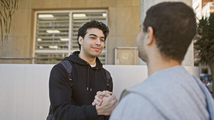 Two hispanic men in casual attire shaking hands on a sunny urban street, displaying a cordial relationship.