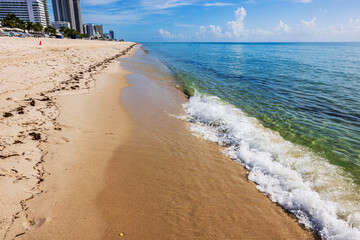 Clear waves gently hitting sandy shoreline on sunny day at Miami Beach with buildings in distance. USA.