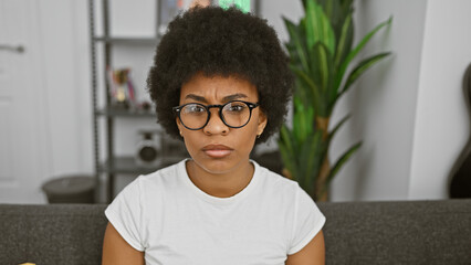 African american woman with curly hair and glasses sits thoughtfully in her modern apartment's living room.