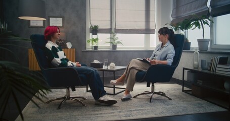 Therapy session in psychological office: teenage girl on consultation. Female psychologist offers glass of water, girl drinks water and shares her feelings. Psychotherapist takes notes in notebook.