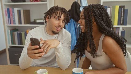 A smiling couple interacts indoors with a smartphone showcasing a casual home setting with books and cosy decor.