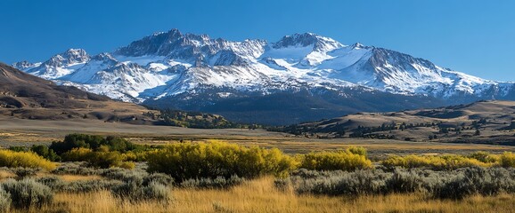 Majestic snow-capped mountain range rising above a valley with golden autumn foliage and a blue sky.