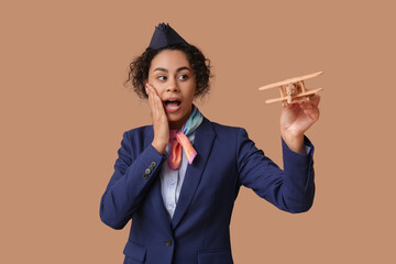 Surprised African-American stewardess with toy plane on beige background