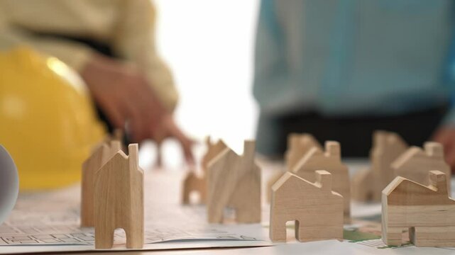 Architect and engineer discuss about building blueprint on table with architectural document, measurement tools scatter around at office, use wooden block for house positioning. Closeup. Delineation.