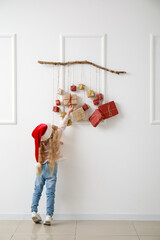 Cute little girl in Santa hat and Christmas advent calendar hanging on light wall