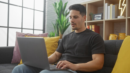 Handsome young hispanic man using laptop comfortably at home in a modern living room setting.