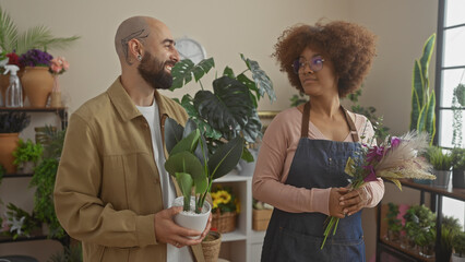 A man and a woman, being florists, smile while holding plants inside a lush flower shop, surrounded by foliage.