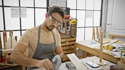 Hispanic carpenter man sanding wood in a well-equipped workshop, wearing safety glasses and casual attire.